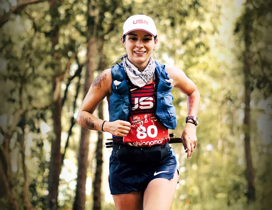 A woman in a white ball cap and a red running jersey jogs through the woods.