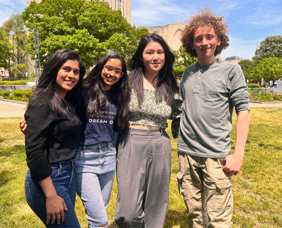 Four students stand with their arms around each other in a grassy plaza.