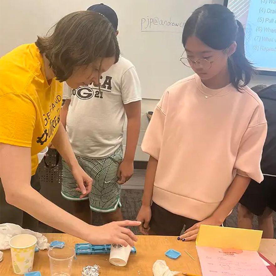 A woman pours liquid from a cup into a mold while another woman watches.