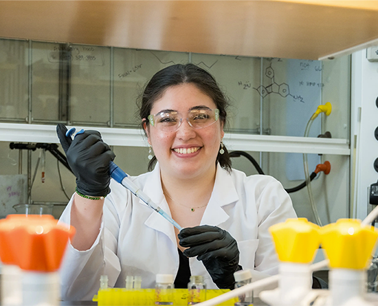 A young woman in a lab coat smiles while holding a syringe.