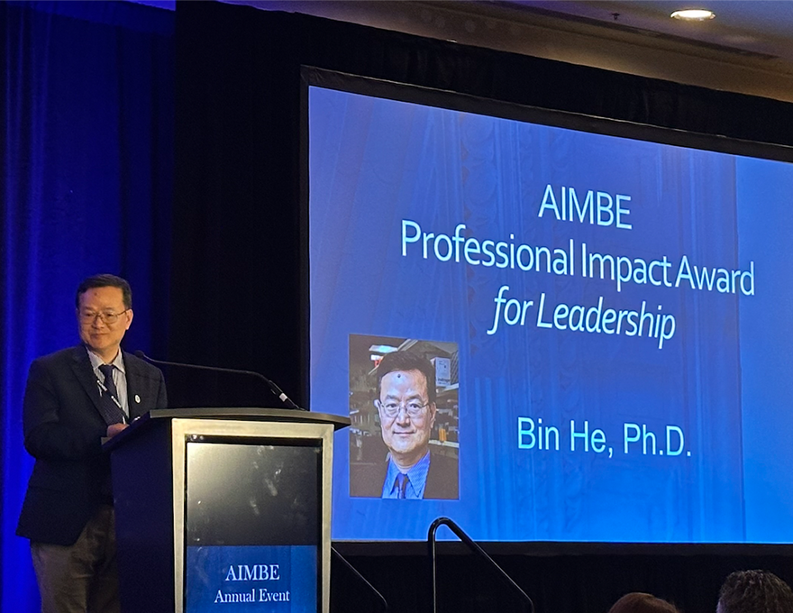 A man in a black suit stands at a podium while a screen to the right says, "A.I.M.B.E. Professional Impact Award for Leadership. Bin He, Ph.D."